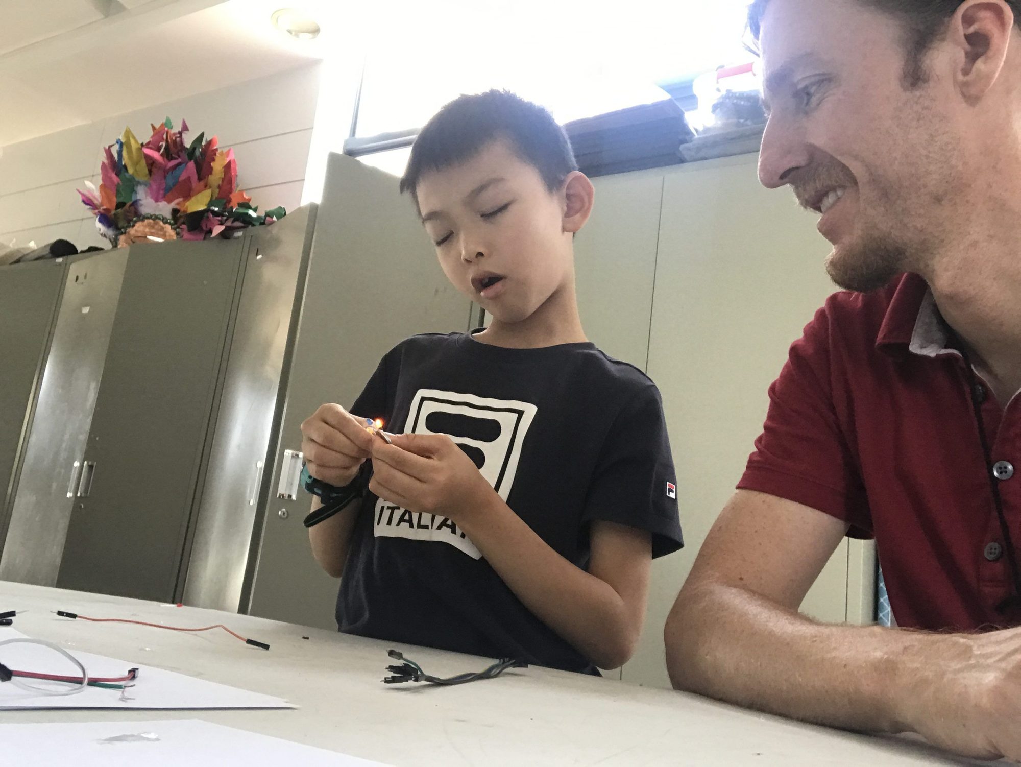 A man in a red shirt shows a boy how to work with a lit LED at a workbench
