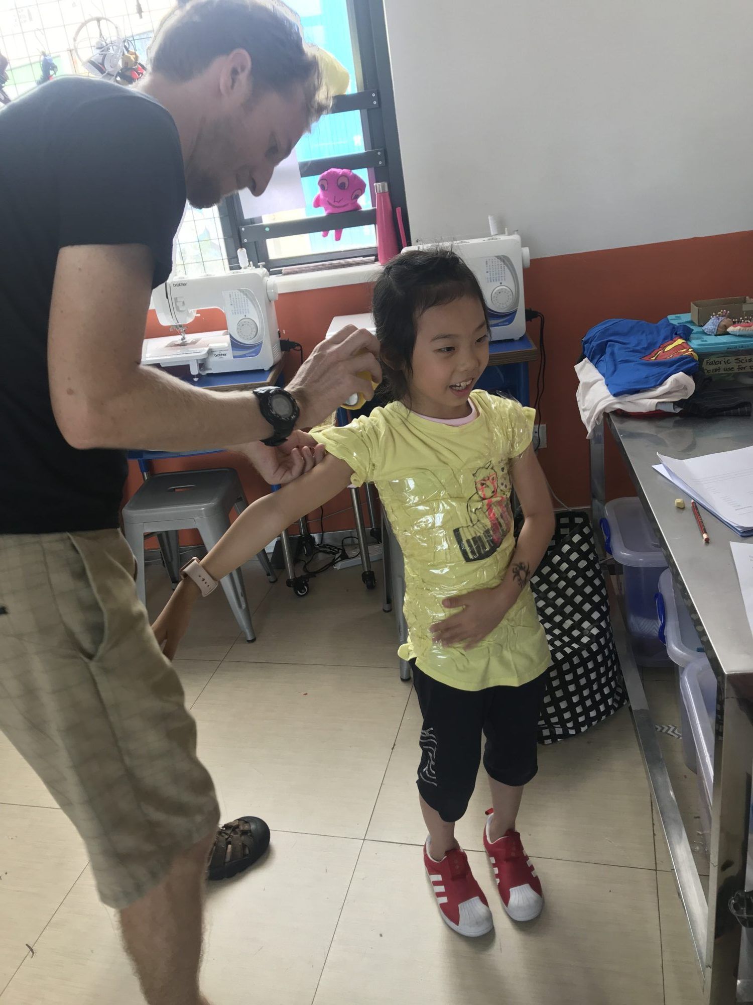 A man attaches a wearable electronics sensor to a smiling girl's arm at a sewing workshop table
