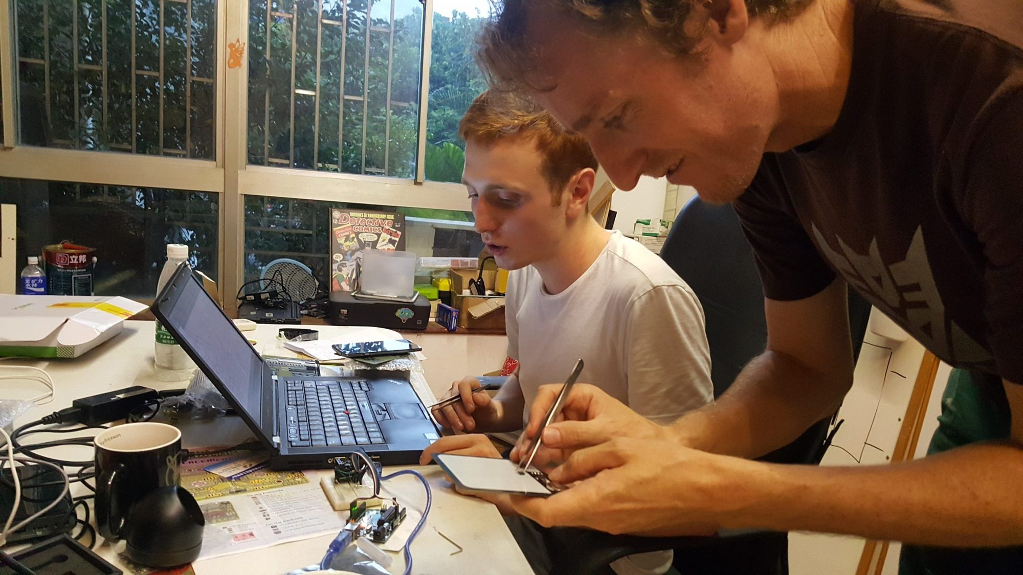 Two young men lean over a laptop and notebook, examining an electronics circuit at a workbench