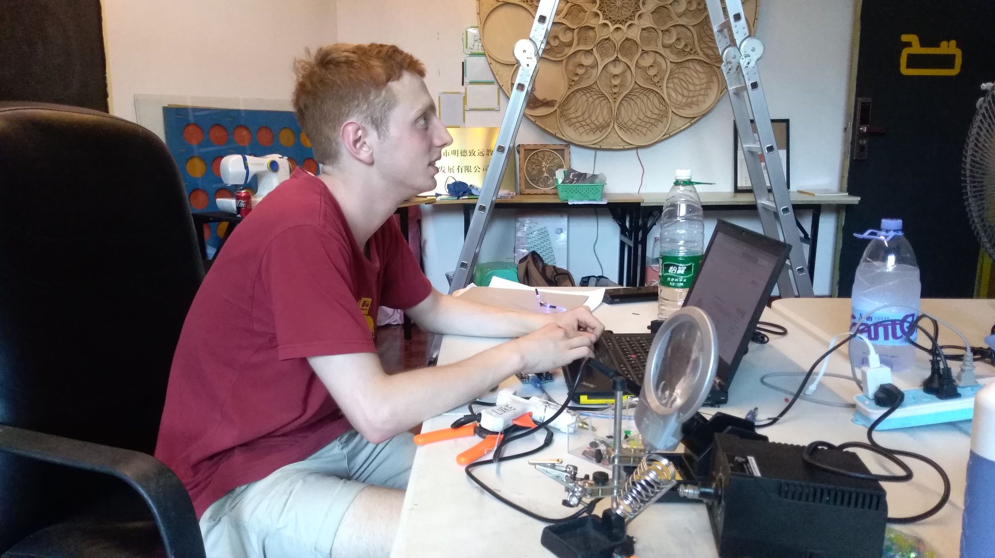 Oliver Child soldering electronics at a workbench beneath a large carved wooden wall sculpture
