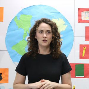 A woman with curly hair speaks in front of a paper cutout of Earth, surrounded by craft displays