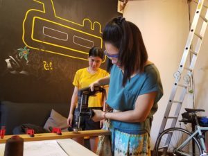 Olivia and another woman assembling a wooden lamp frame with a drill in a workshop
