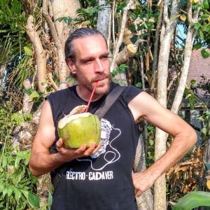 Marc Dusseiller drinking from a fresh coconut through a straw among green foliage