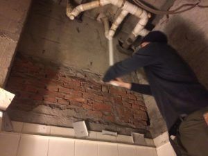 Person kneeling to inspect an exposed pipe under a broken tile floor