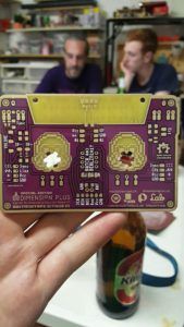 A hand holds a purple circuit board while two people talk in the background