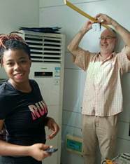 A man holds a giant yellow pencil above a laughing woman in a workshop