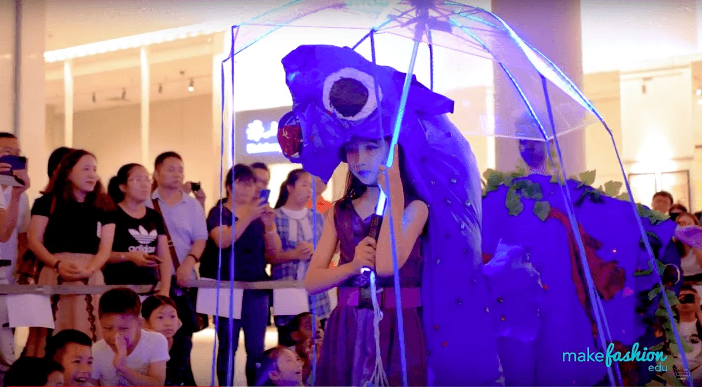 Model in a glowing indigo costume with umbrella at the Shenzhen MakeFashion Edu runway show