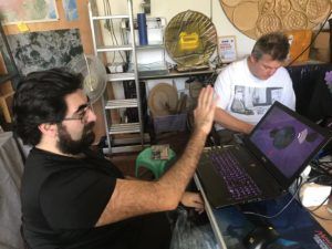 Two men laughing at a laptop, one raising a hand