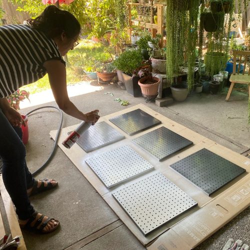 Mobile Makerspace chapter 9: a volunteer spray-painting pegboard panels in a plant-filled driveway