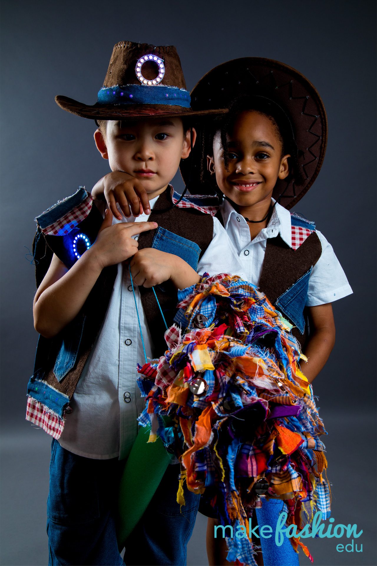 Two student models in cowboy hats and patchwork vests, one holding colorful fabric horse prop
