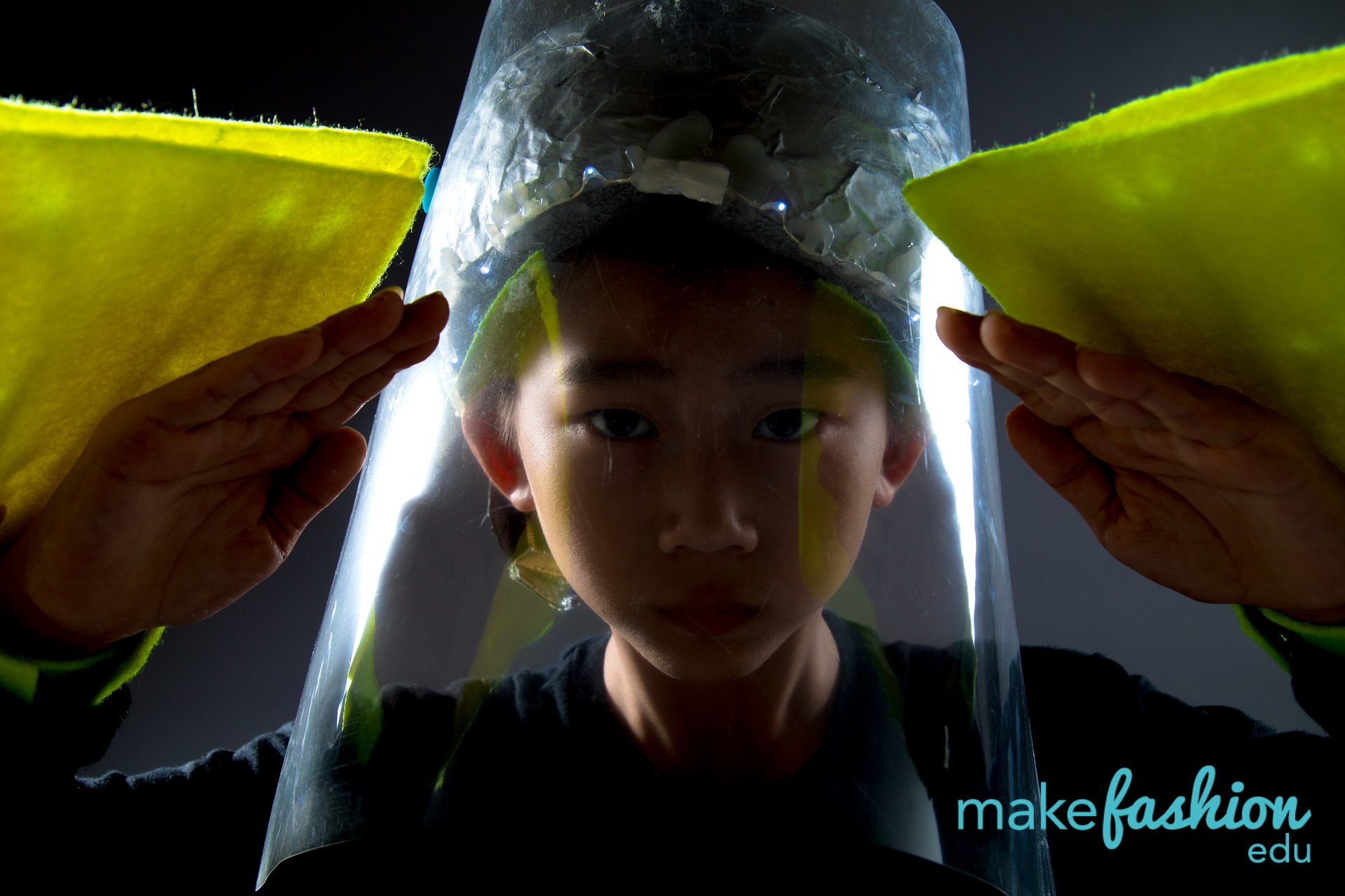 Student in clear illuminated veil with small white LEDs, hands holding yellow felt wing panels