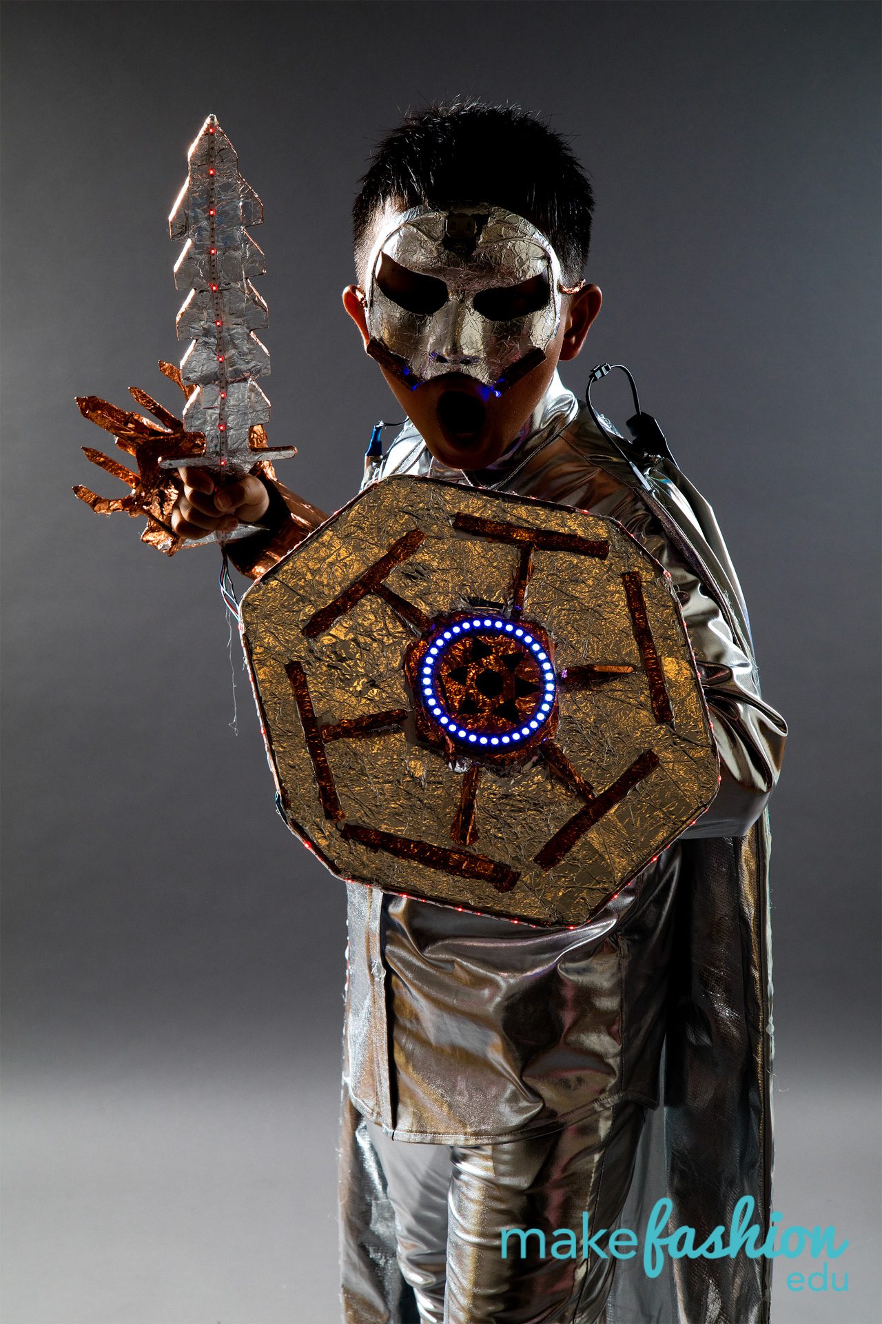 Student model in silver mask holding foil sword, gold shield with blue LED ring center