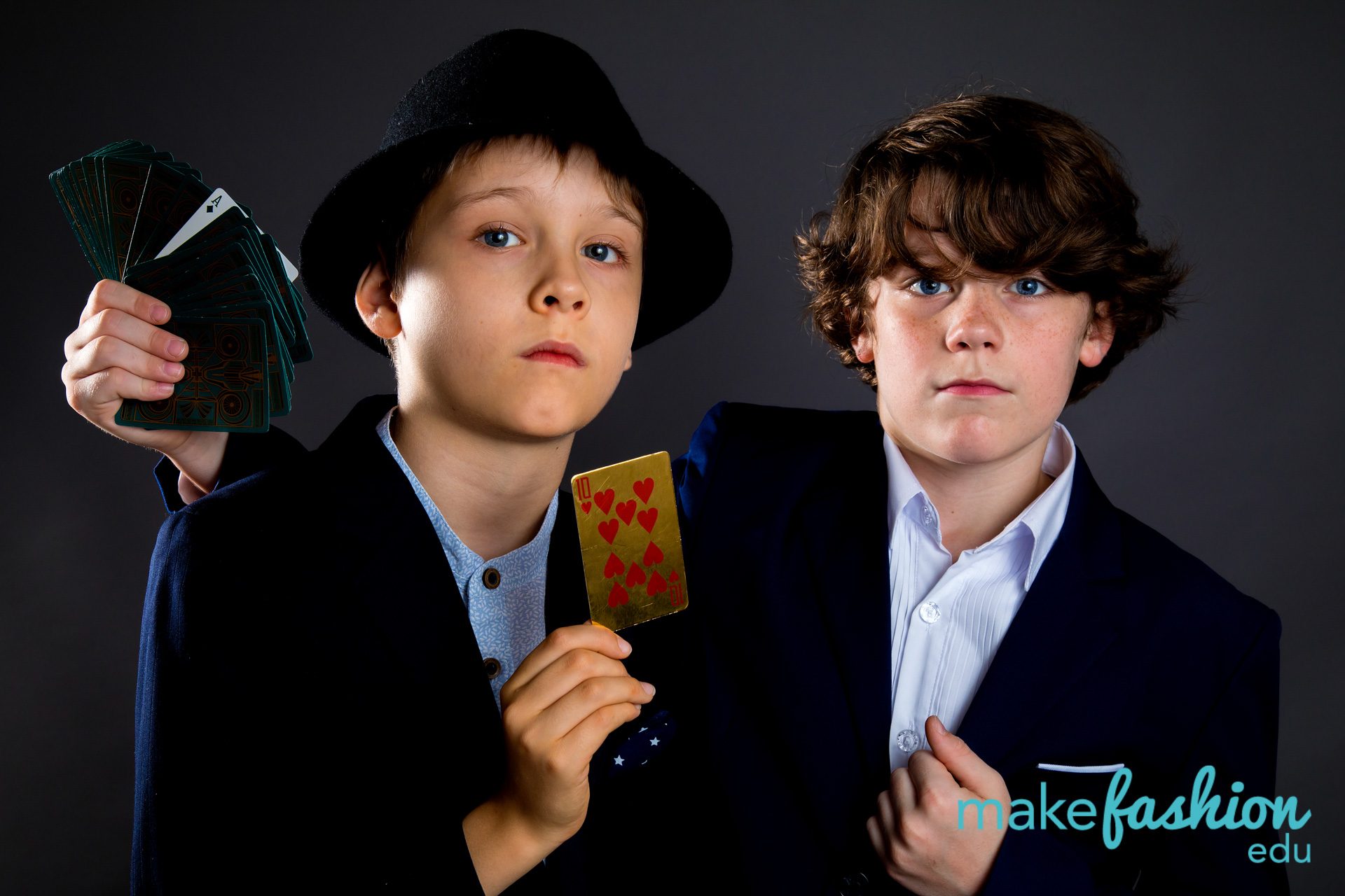 Two student models in navy blazers holding playing cards, one wearing a black hat