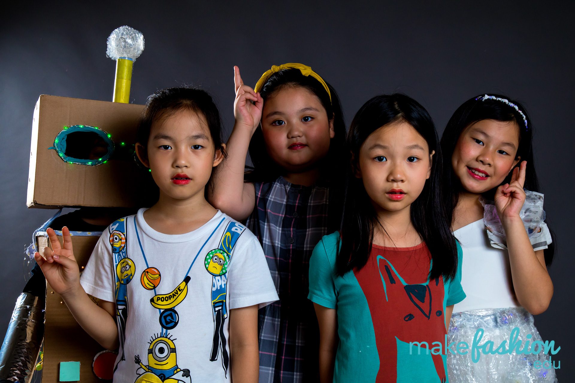 Group of four student designers with cardboard robot head featuring LED eye and various festive accessories