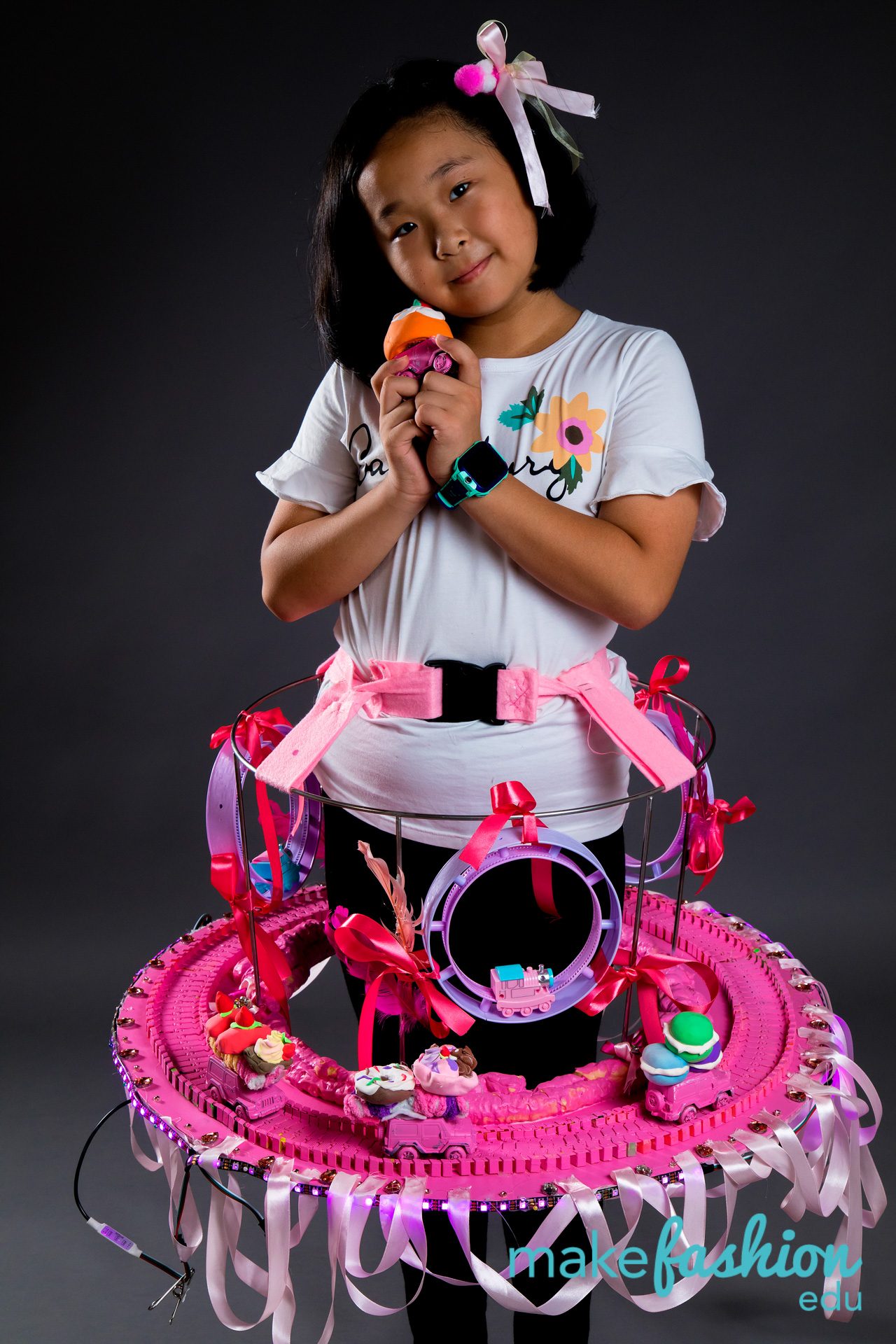Student in white shirt wearing large pink sculptural skirt with toy vehicles and lights