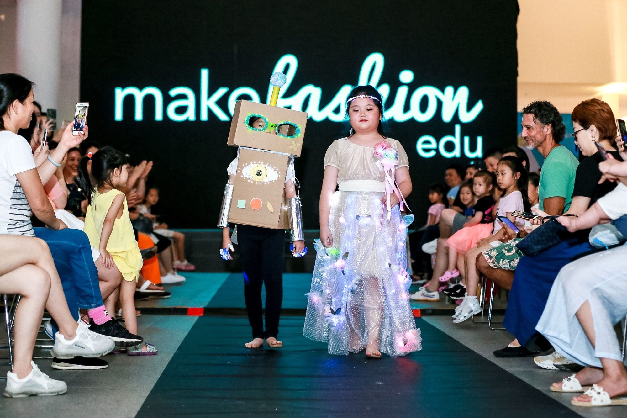 Runway show with student model in white illuminated tulle dress and cardboard robot head with LED eyes