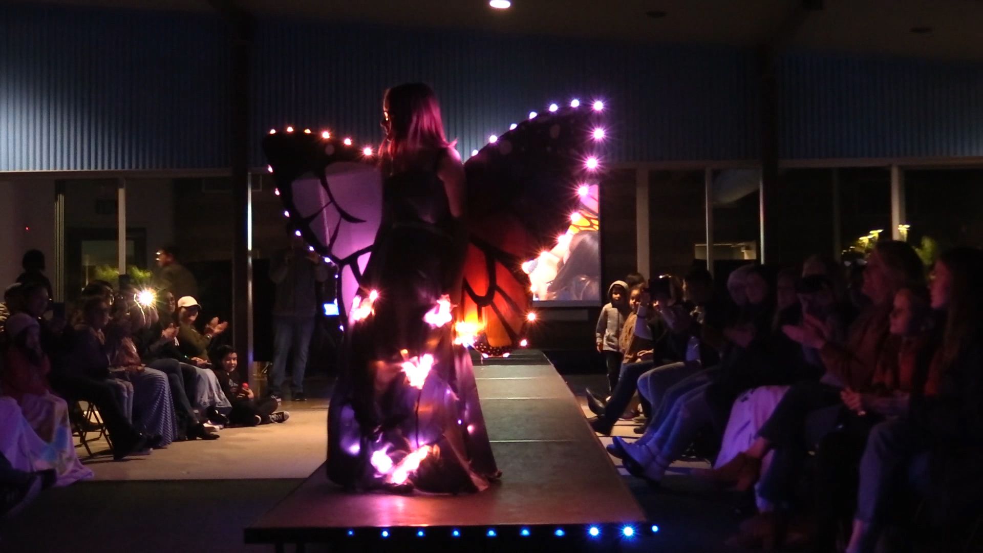 Student on runway with large illuminated butterfly wings with colorful LEDs