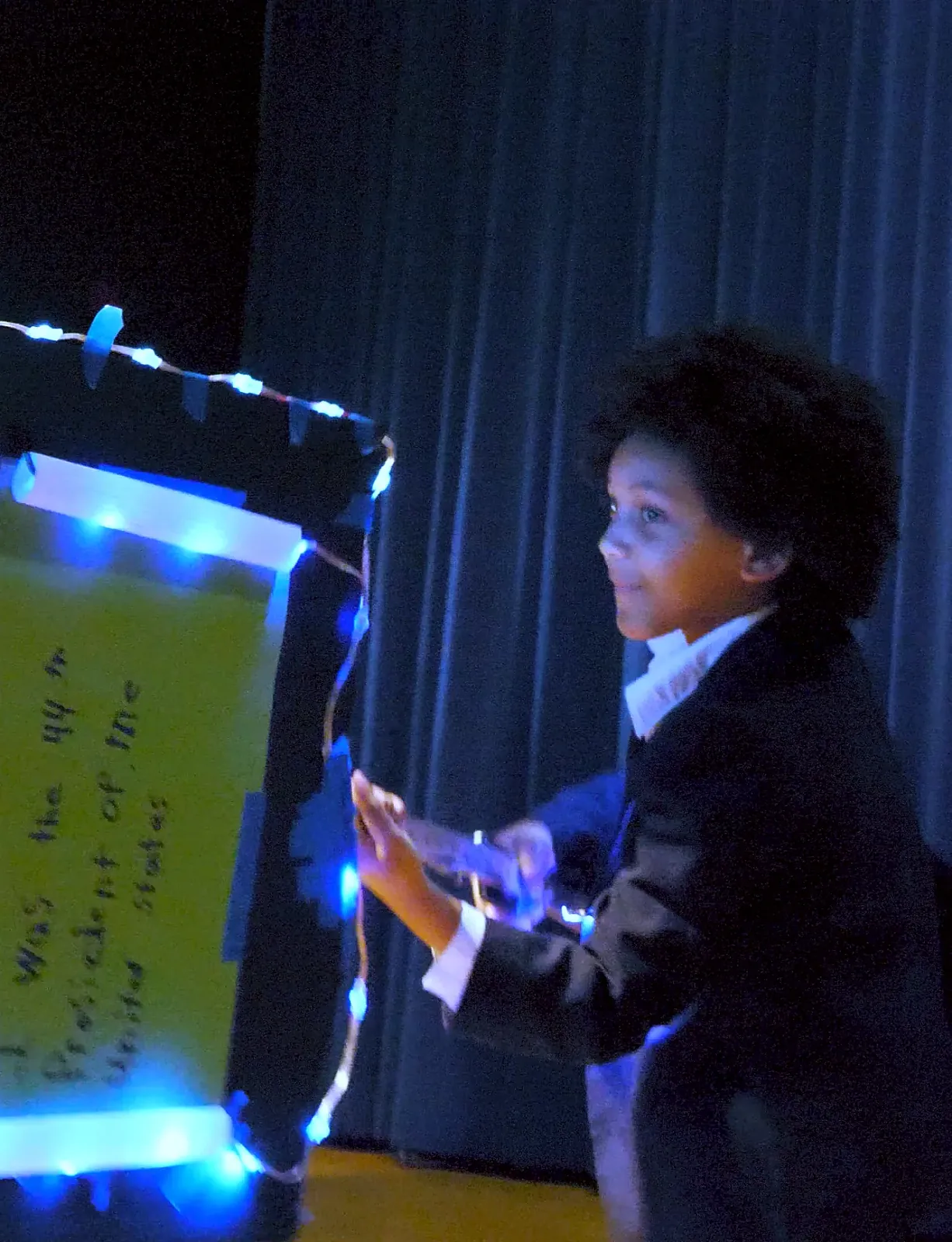 Student as Barack Obama on stage beside a scroll framed in blue LEDs that reads "was the 44th president of the United States"