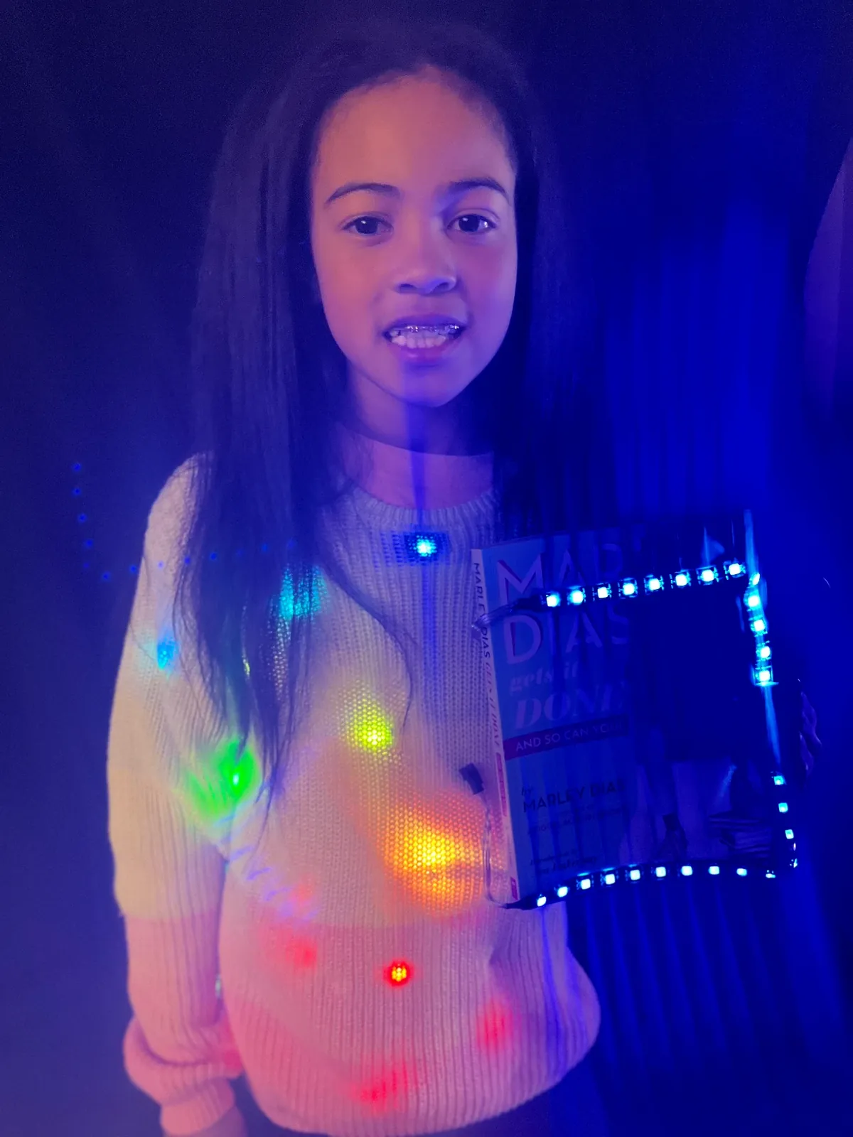Student as Marley Dias in a cream sweater lit with rainbow LEDs, holding her book Marley Dias Gets It Done framed in blue LED strips