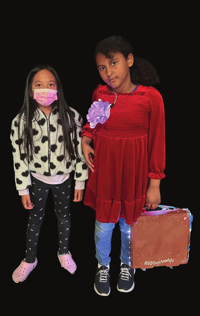 Two students as Linda Brown and a classmate, one in a red velvet dress with a flower clip carrying a light-up suitcase