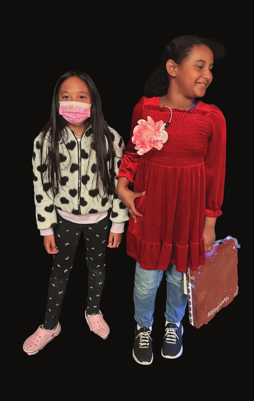 Student as Linda Brown in a red velvet dress beside a classmate in a heart-print sweater, a suitcase at her side