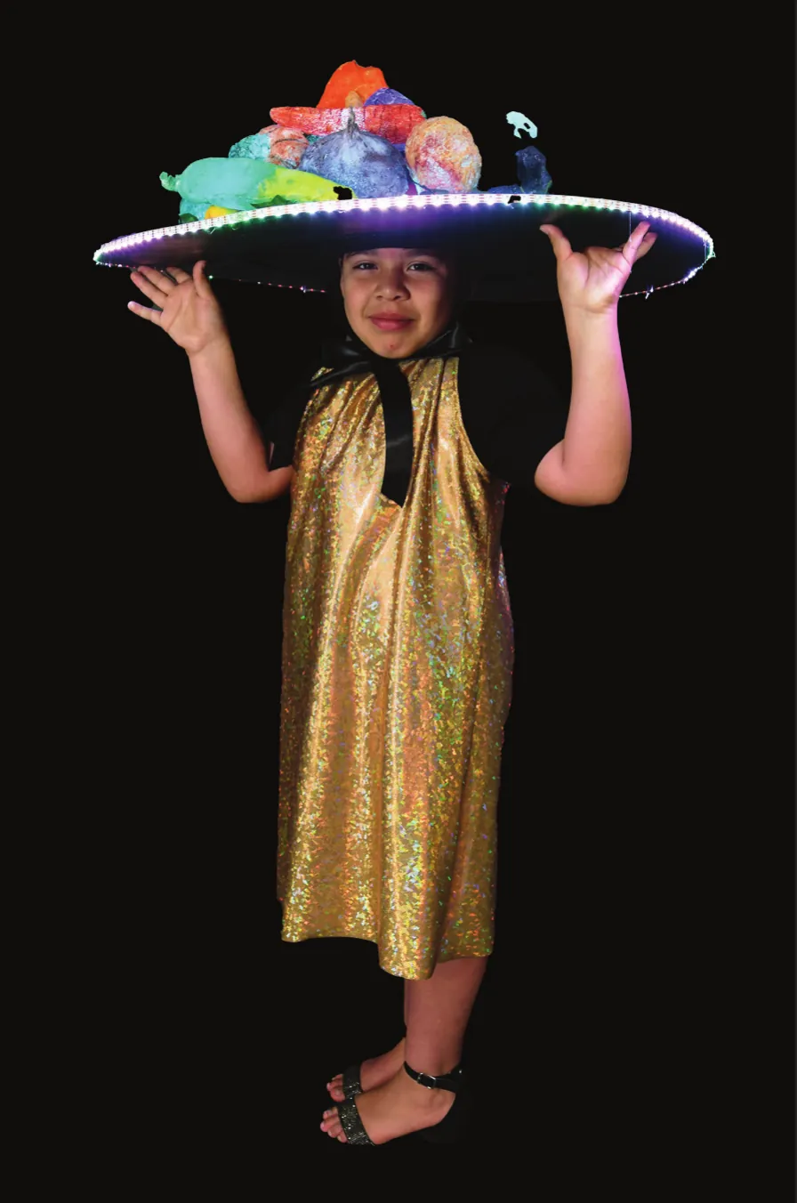 Vegging Out: student in a gold dress holding up a wide hat piled with colorful felt vegetables